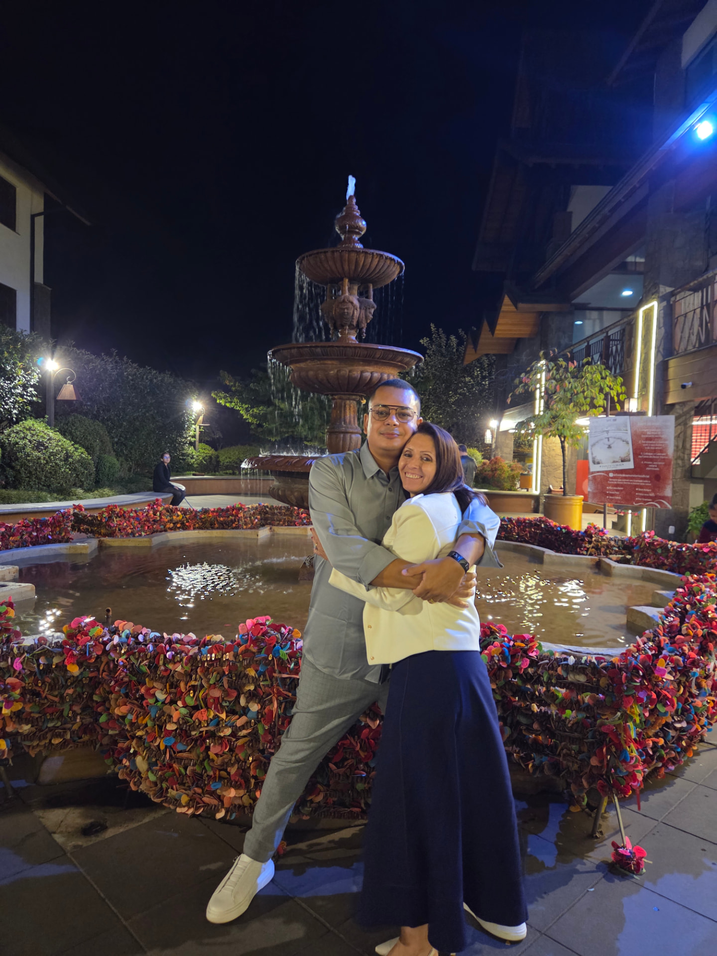 Renato Medeiros abraçado com sua esposa, Marcia Coelho, em frente a uma fonte iluminada à noite